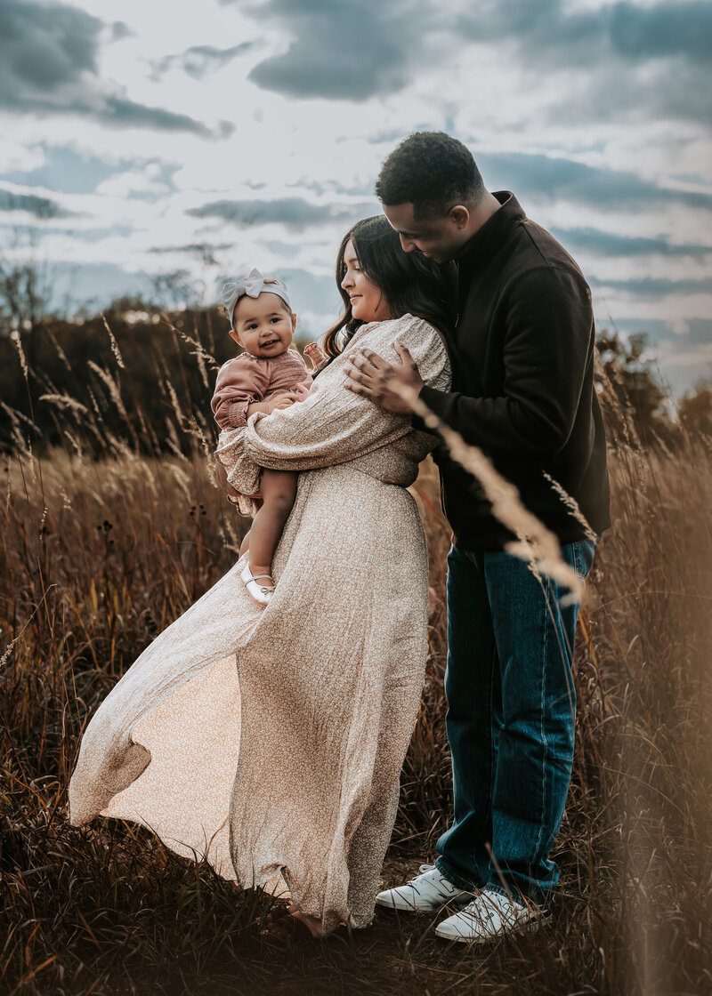 A young family sits in the grass next to a tree and snuggles and laughs together