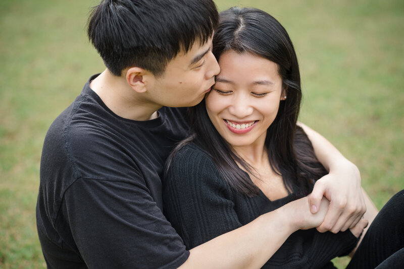 Romantic proposal moment at Daniel Stowe Botanical Garden, captured by Charlotte proposal photographer — man kisses smiling woman’s forehead as they embrace on the lawn.