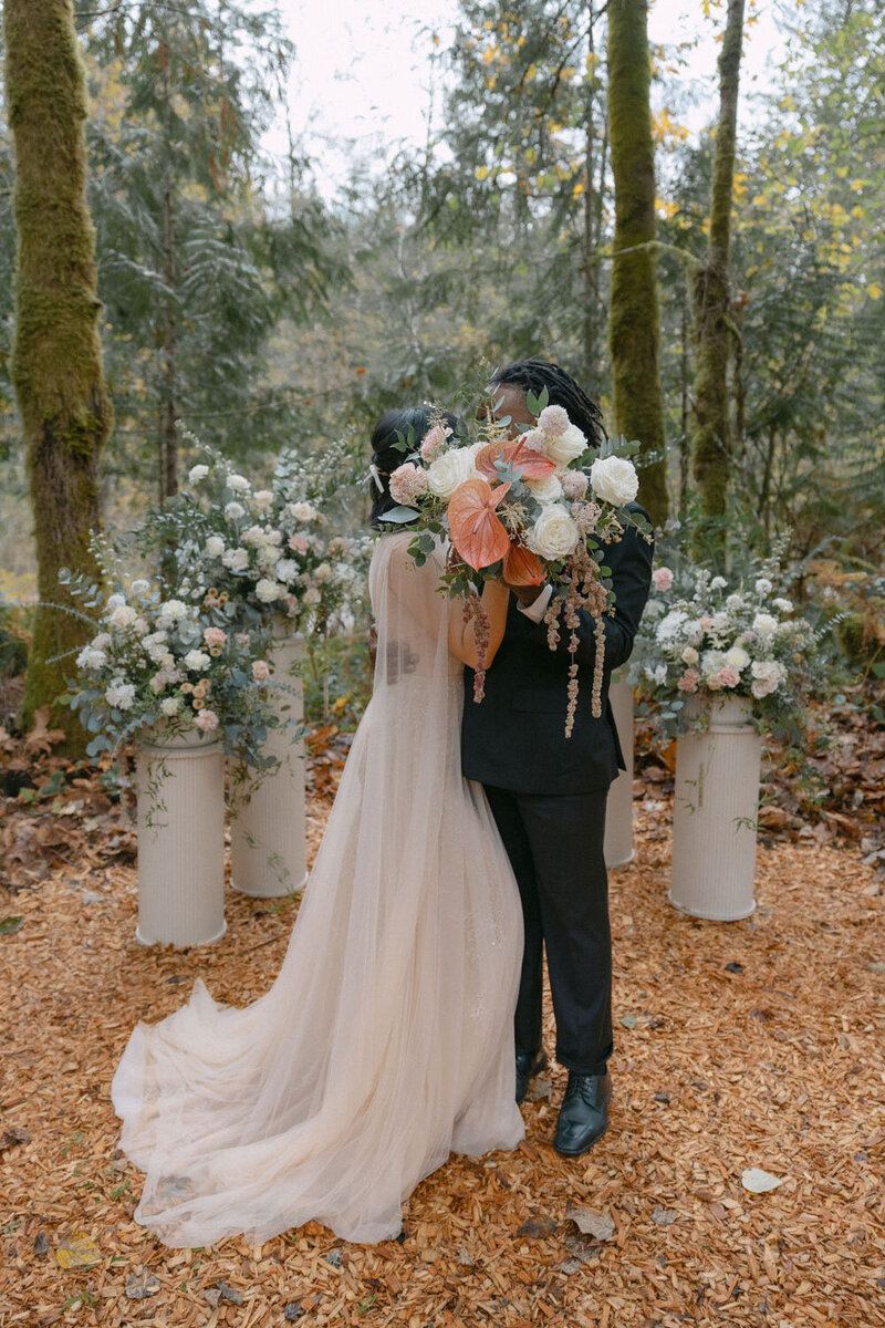 Couple sharing a kiss behind a lush wedding bouquet in the forest