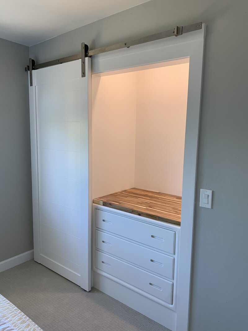 A white barn door is open showing a closet space with a white built in dresser and wood top. There is a light shining inside above the dresser with an open white space.
