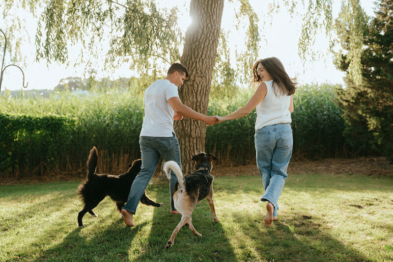 couple running in grass with dogs during at home engagement photos, captured by Elsie Goodman, an NYC engagement and couples photographer