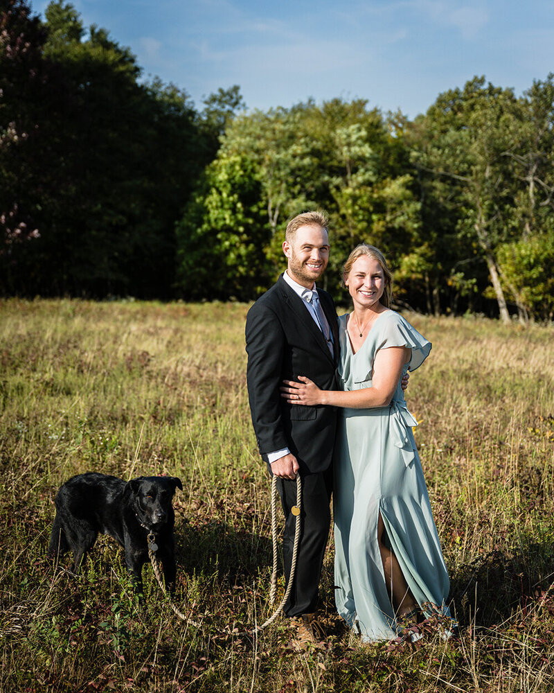 A bride and groom stand side-by-side for a photo with their dog, who is leashed, in a field in Shenandoah National Park.