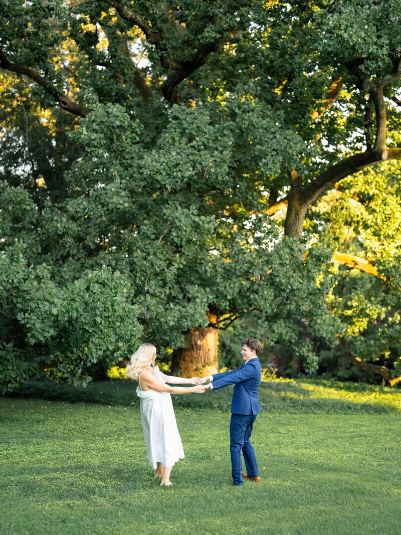 A couple dances joyfully on a grassy field, surrounded by lush green trees. The woman wears a white dress, and the man is in a blue suit. Sunlight filters through the branches, creating a serene and romantic atmosphere.