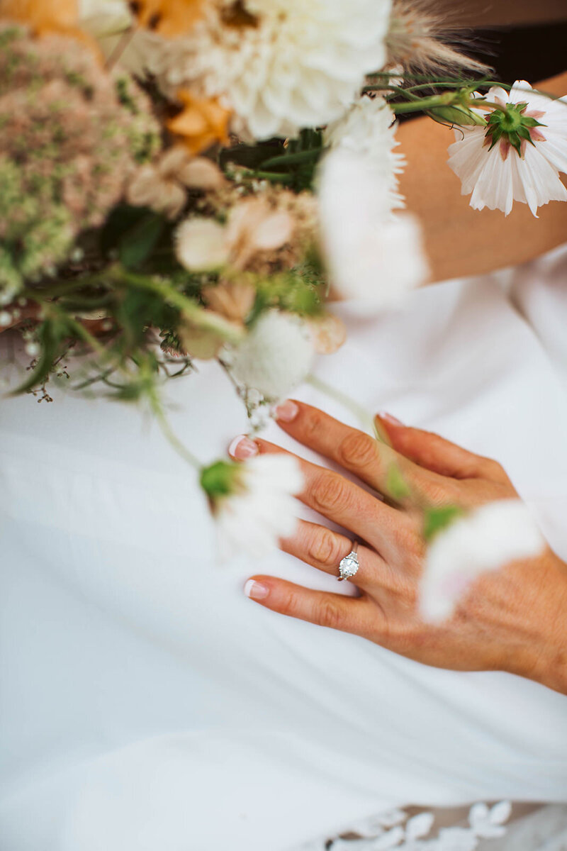 Close-up of bride’s hand with engagement ring resting on her gown, holding a bouquet of wildflowers at a Maine wedding.