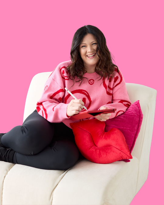 Emily, branding expert, sitting on a white soft chair in a pink sweater with her iPad designing brand assets.