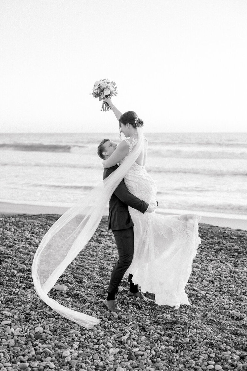 black and white photo of a groom lifting his bride and twirling her on the beach