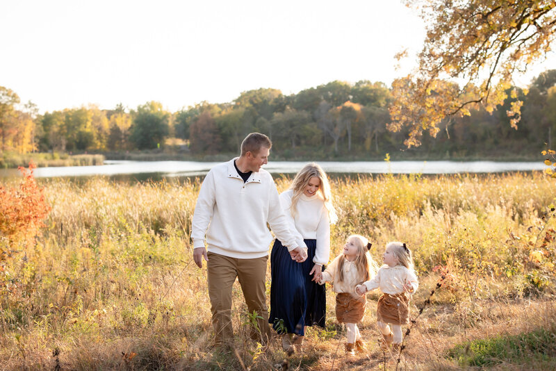 a family of 4 wearing white and neutral clothing hold hands a they walk through a grassy field at golden hour with a lake in the background.