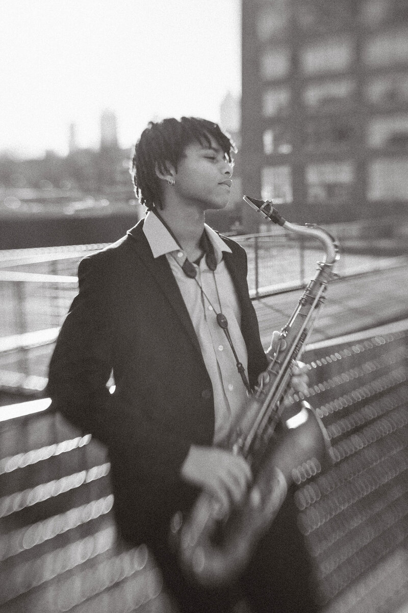A senior boy leaning against a parking garage wall holding a saxophone. 