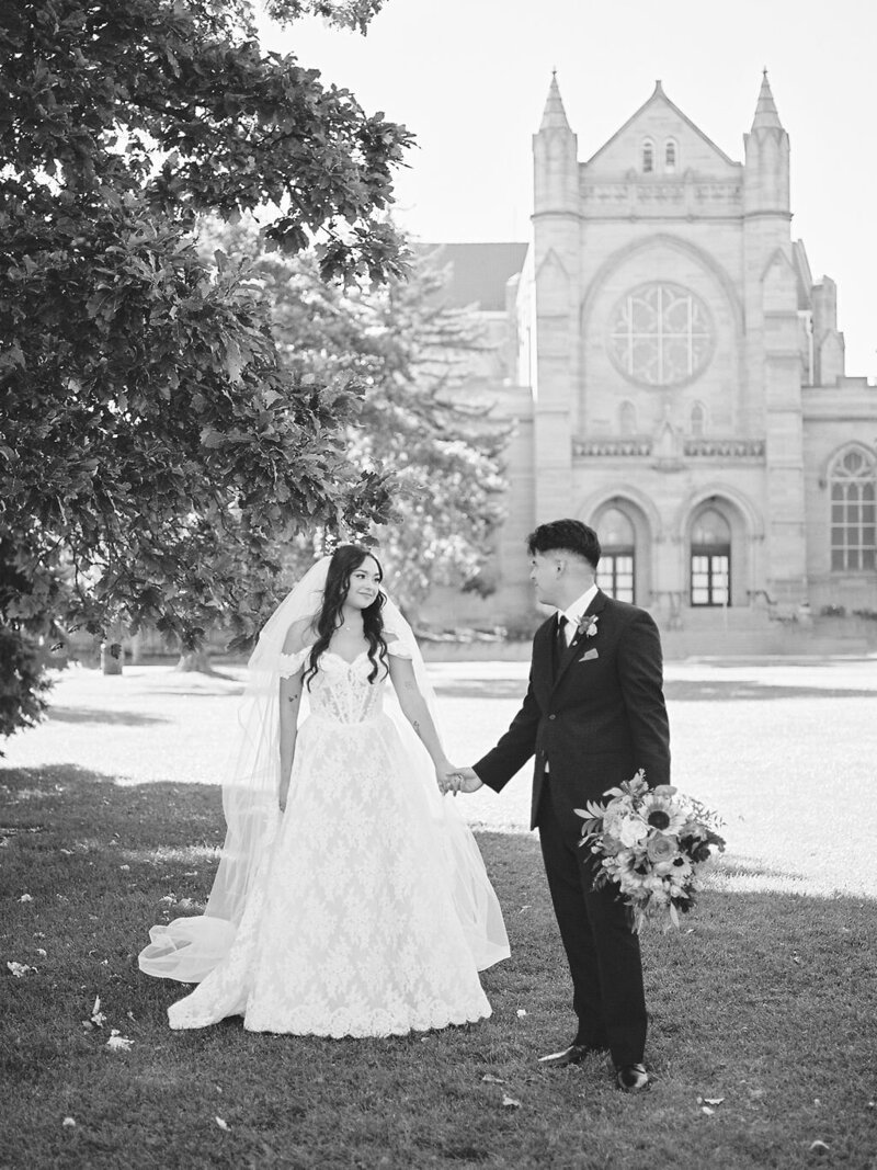 A bride and groom hold hands, looking at each other in front of the Catholic Church they got married in, located in Denver, CO.