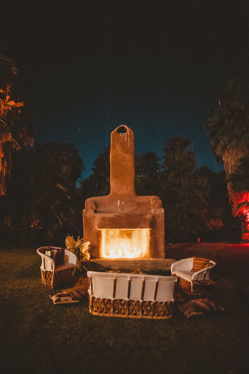 Nigh time photo of a stone outdoor fireplace with canvas and wood chairs around it.