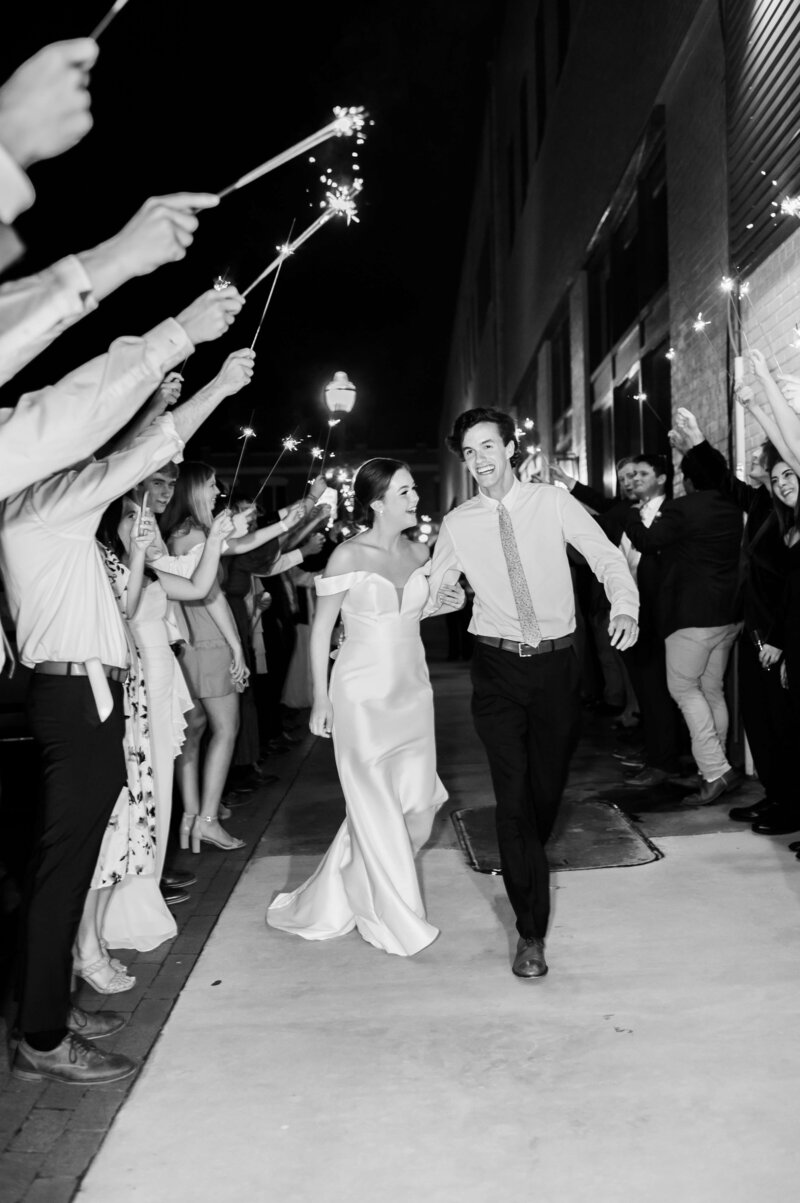 A bride and groom walk hand in hand outside at night, smiling as guests on both sides hold sparklers and cheer, creating a festive tunnel for their wedding celebration. The scene is in black and white.