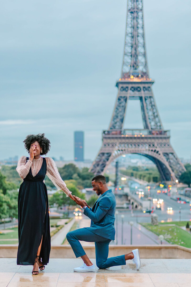 Man kneeling ready to proposal by Eiffel Tower as women shields her eyes during surprise proposal in Paris