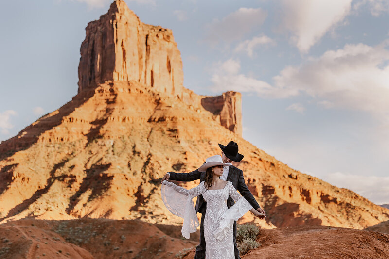 A groom stands behind his bride as they are hand in hand. She is wearing a beautiful lace dress with oversized sleeves, and they are both wearing cowboy hats. They are celebrating their elopement in Moab.