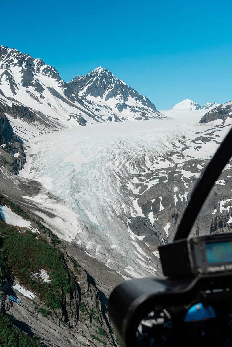 Rugged snow capped mountains are viewable out of the cockpit during private helicopter flightseeing tour headed to a private dogsledding experience for a luxury elopement package near Bear Glacier in Seward.  There is a snowy, crevasse filled glacier spilling out from the mountains at the top of the frame and running down valley between the rugged peaks.  Helicopter instruments are viewable in the lower right corner of the frame. 