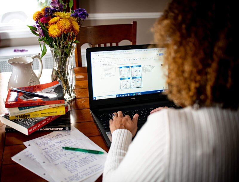 Woman with back to camera typing on a computer at the dining room table with flowers.