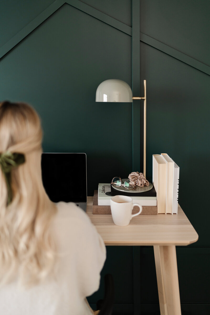 Blonde Frau sitzt am Schreibtisch vor ihr ein Laptop und eine Kaffeeteasse. Die Wand ist dunkelgrün