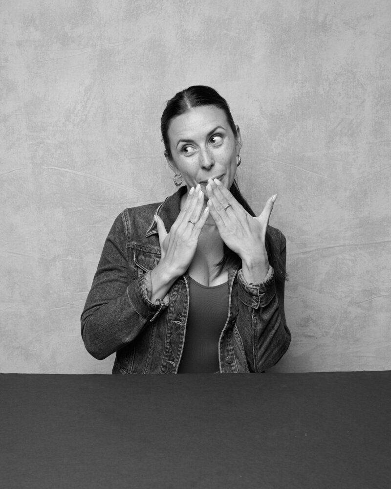 Black and white portrait of a woman playfully covering her mouth with both hands, her eyes glancing sideways with a mischievous smile. She wears a denim jacket over a fitted top, exuding confidence and authenticity. A fun, unfiltered moment from 'THIS IS ME – A Photography Experience by Katie Campbell' in Atlanta, GA, celebrating self-expression and individuality.
