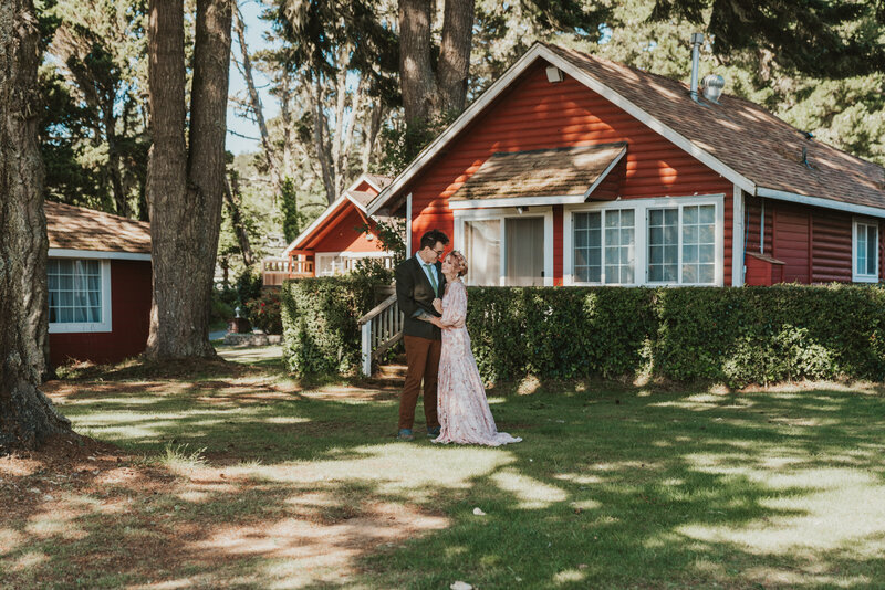 Bride wearing a pink wedding dress holding groom wearing a green suit jacket and tie looking at bride in front of red cabin by California elopement photographer Kasey Mantiply