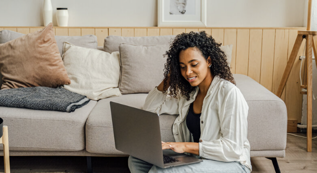 Girl sitting on the ground next to couch with her laptop on her lap. 