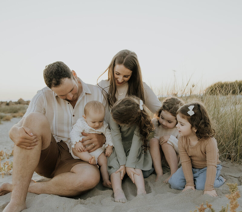 Family on the beach, mum and dad with four young children gathered on their laps and around them.  Photo is light and airy, three older children and their parents are smiling and looking at the youngest child, a baby.  