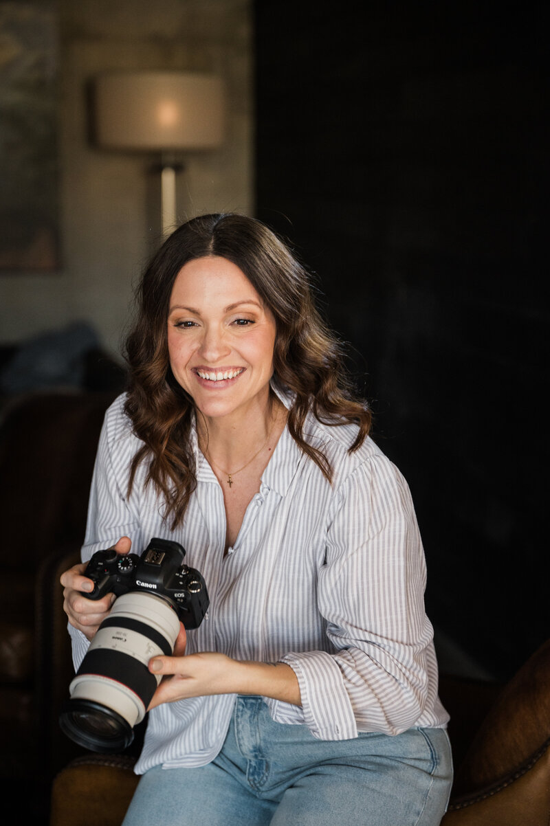 A smiling woman, participating in a photography mentoring program, holding a camera, seated in a cozy room with soft lighting.