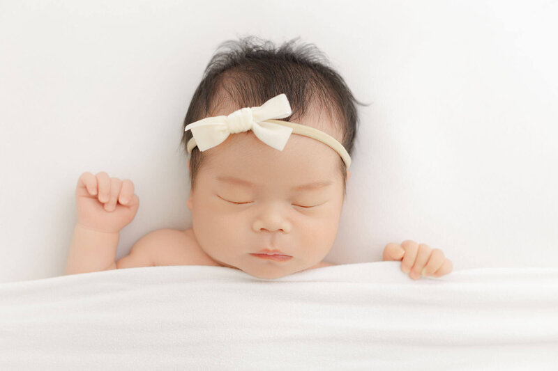 Baby girl with ivory bow sleeping under a white blanket. One hand is up by head and the other hand is holding onto the edge of the blanket.