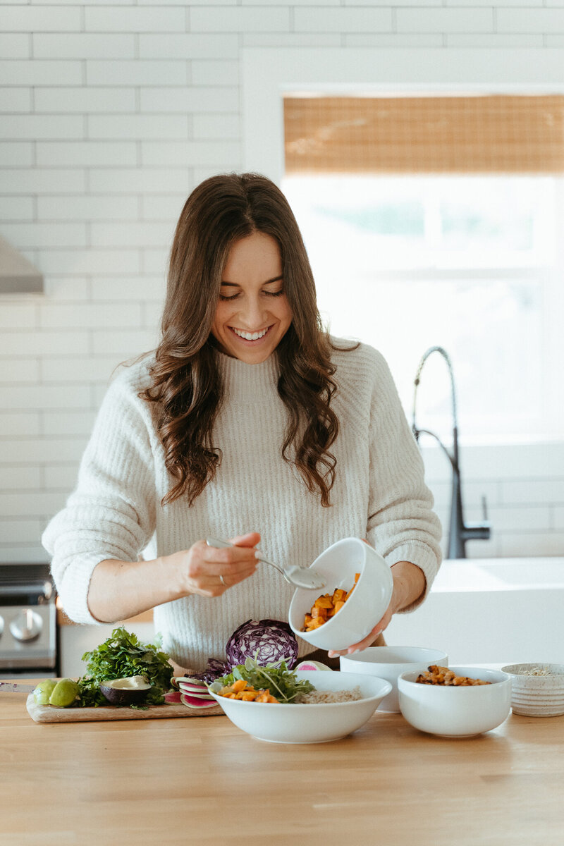 Woman with brown hair and a tan sweater seasoning food in the kitchen