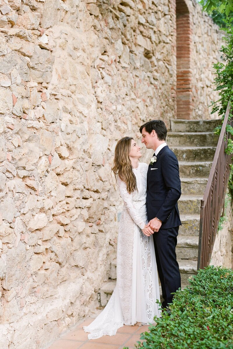 Bride and groom embrace at base of stairs in front of a stone wall