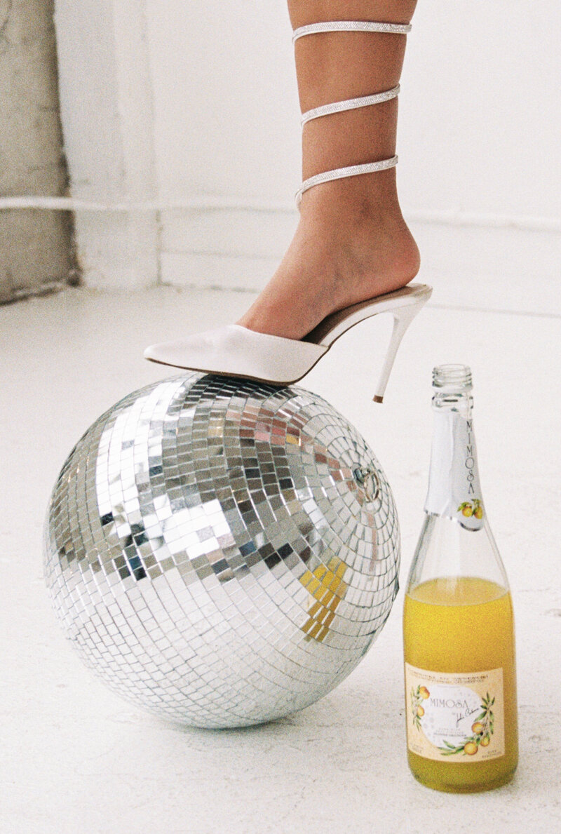 White heel sitting on a disco ball next to a bottle of mimosa