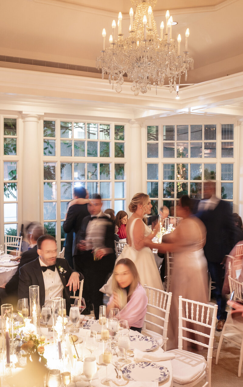 A warmly lit, elegant dining room with guests in formal attire. People are seated at round tables adorned with floral centerpieces, under a chandelier. Large windows allow in natural light. Some guests are standing and conversing.