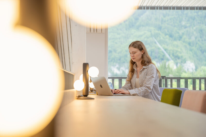Woman working on her laptop with stunning mountain views behind her, enjoying the perfect blend of productivity and nature at The Campus Alps.com for private getaways and company retreats.