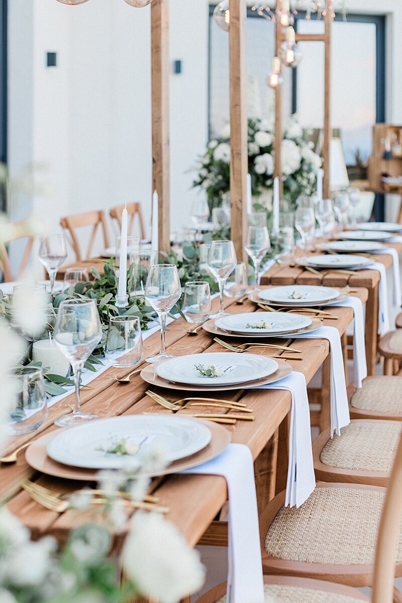 Minimalist outdoor wedding tablescape with wooden tables, neutral linens, gold cutlery, and greenery table runners for a modern elegant celebration