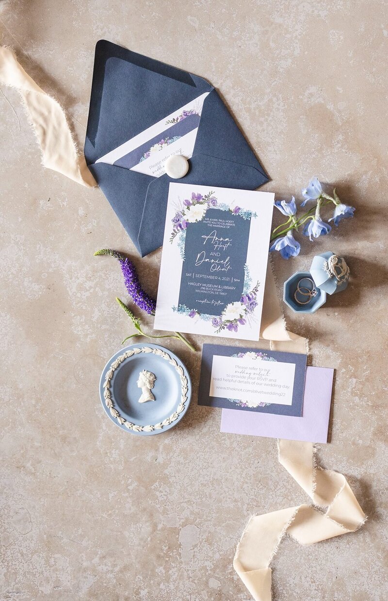 Flatley of invitation suite, in periwinkle, light lavender on a beige flatlay mats, blue dishes and wedding ring.