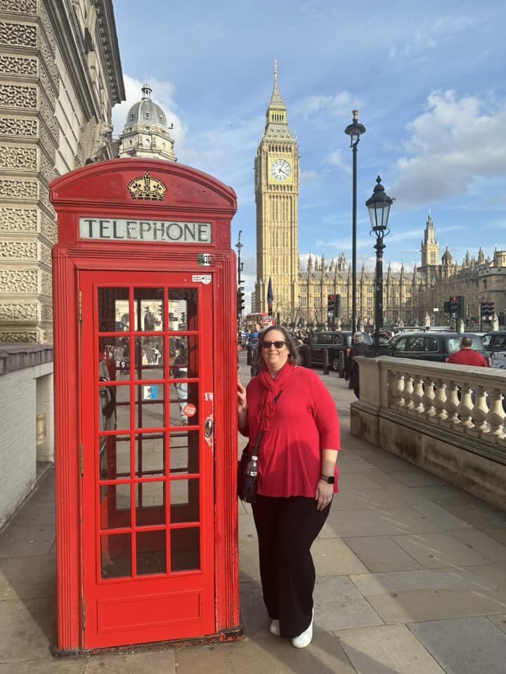 A woman, in a red shirt, is leaning against a iconic London red telephone booth with Big Ben behind her in the distance.