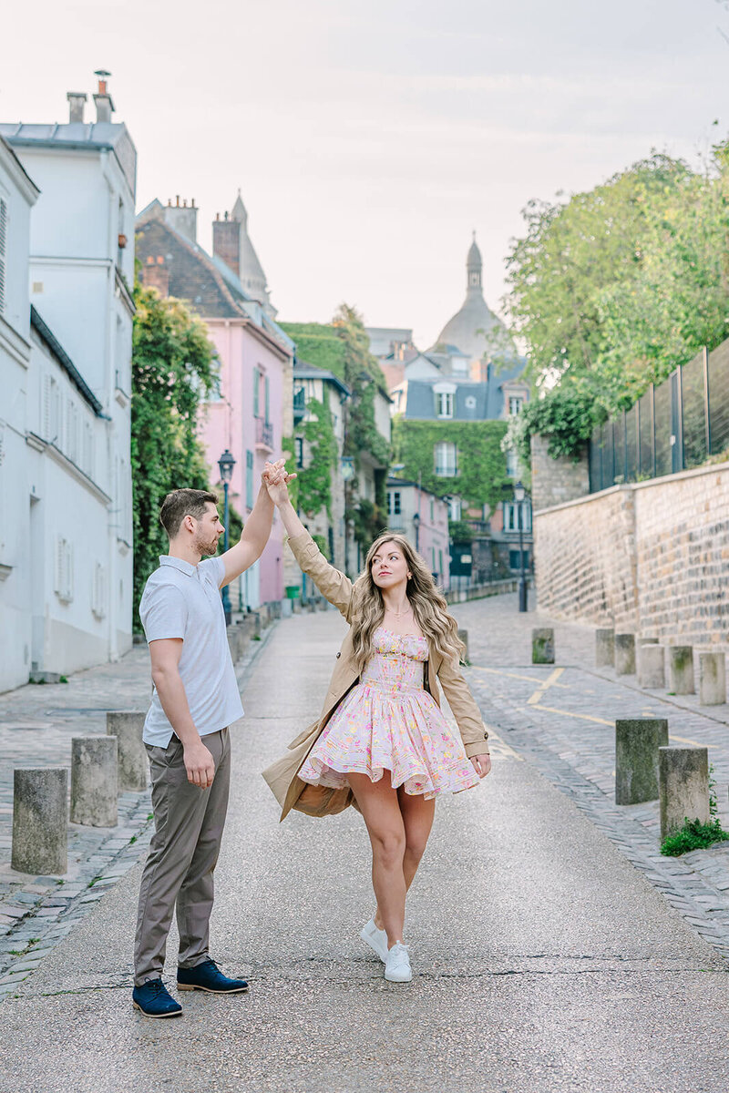 Man and woman walking hand in hand down the street near Sacré-Cœur and rue de l’Abreuvoir in Montmartre, with the man playfully about to spin the woman during a romantic Paris couples photoshoot.