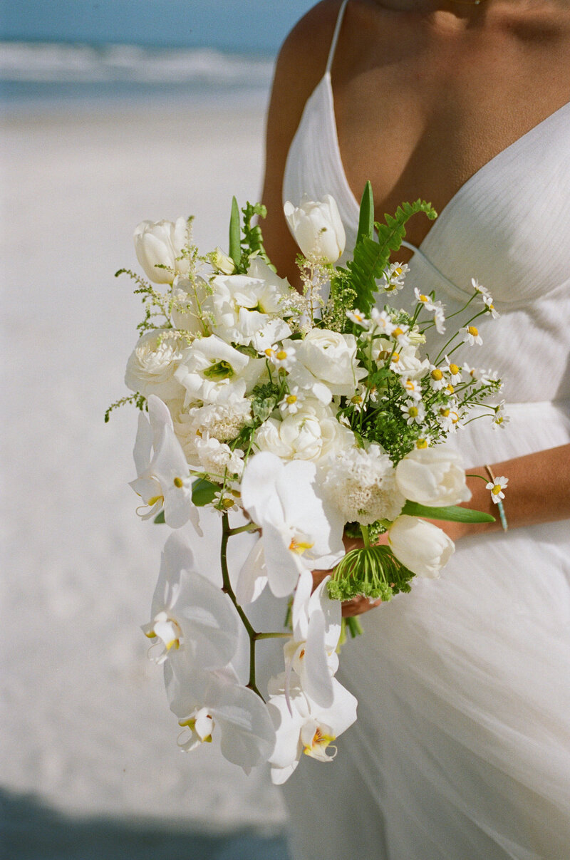 Close-up of a bride holding a cascading white bouquet with orchids, tulips, and wildflowers on a beach. Captured on 35mm film by The Detablans. 