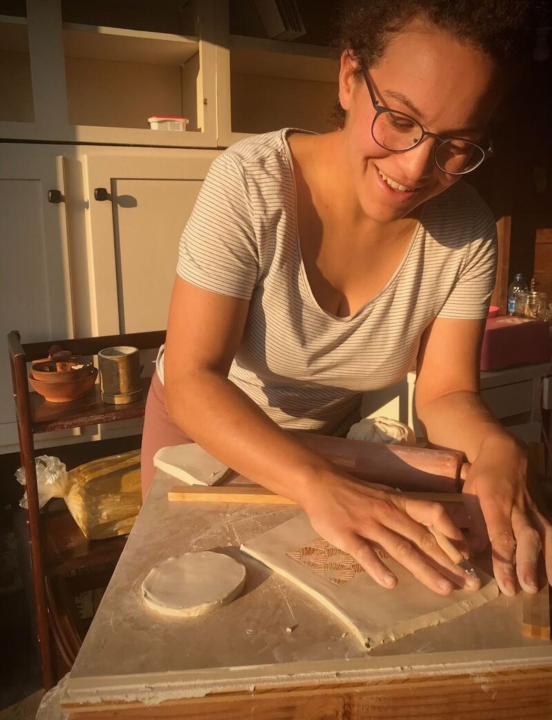 Women in golden light, wearing dark rimmed glasses and striped shirt, smiling down at her hands as she rolls out clay