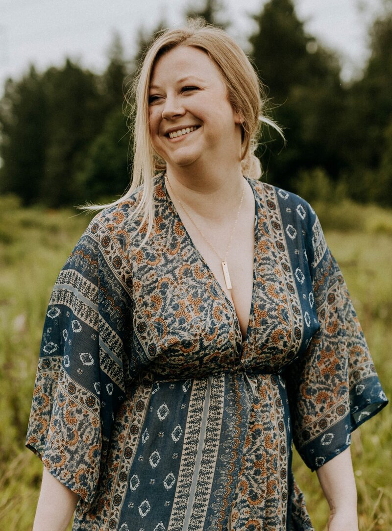 Jaymie Sutherland, Spiritual Healer, Activator, & Expander, standing with a bridge in the background, looking over to the left with a hands clasped in front of her & a soft smile on her face.