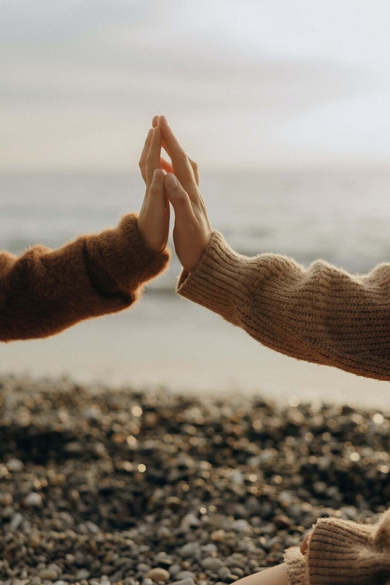 Zwei Hände, die sich vor der Kulisse eines kieseligen Strandes berühren: Verbindung und Unterstützung auf dem Weg zu persönlichem Wachstum