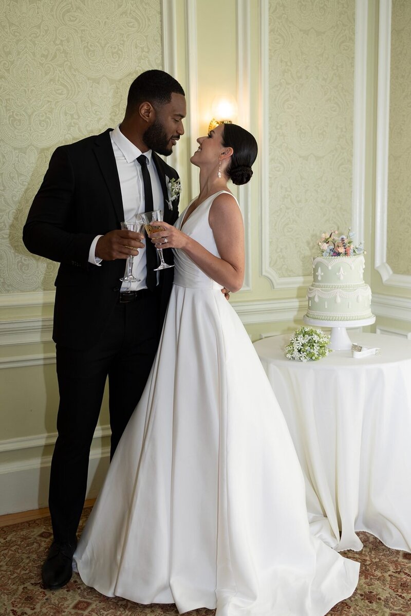 Inter-racial couple holding champagne glasses during a reception moment with a beautiful green cake behind them.