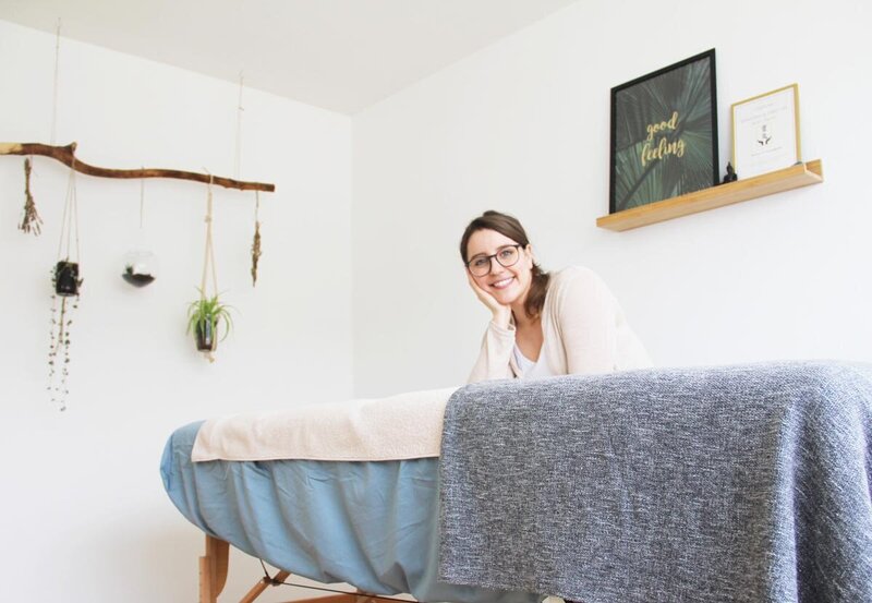 Picture of Nathalie in her practice. Sitting by massage table, smiling