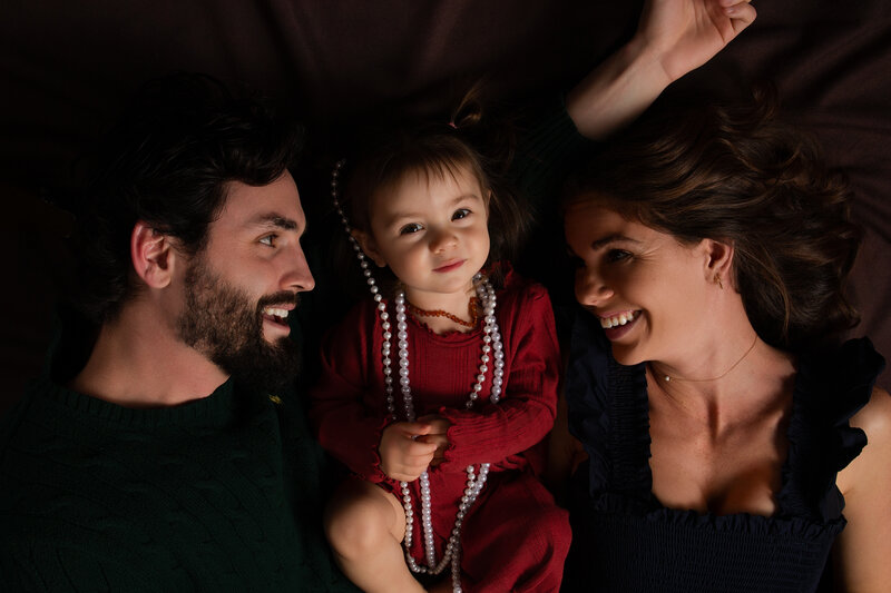 dad, daughter and mom laying on floor everyone looking at the toddler, while she is looking at the camera smirking while wearing pearls. All smiles in Charlotte, NC