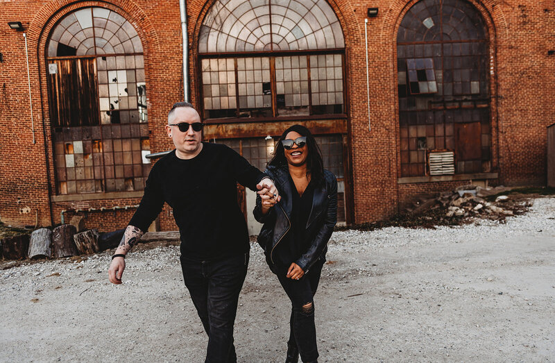 Couples photography by Baltimore wedding photographers with man and woman holding hands and walking next to an old abandoned warehouse with the large windows and brick
