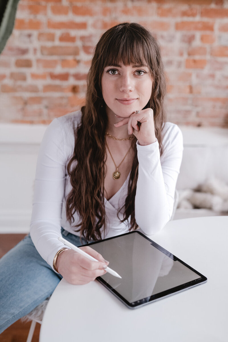 The founder and designer of WildHive Studio is dressing in a white, crossbody top and jeans while looking at the camera. She is drawing on her iPad Pro using a white iPad Pencil.
