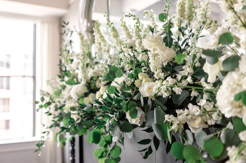 A lush floral arrangement featuring white roses, chrysanthemums, and delphiniums alongside green eucalyptus leaves. The arrangement sits against a dark background, with natural light streaming in from a nearby window.