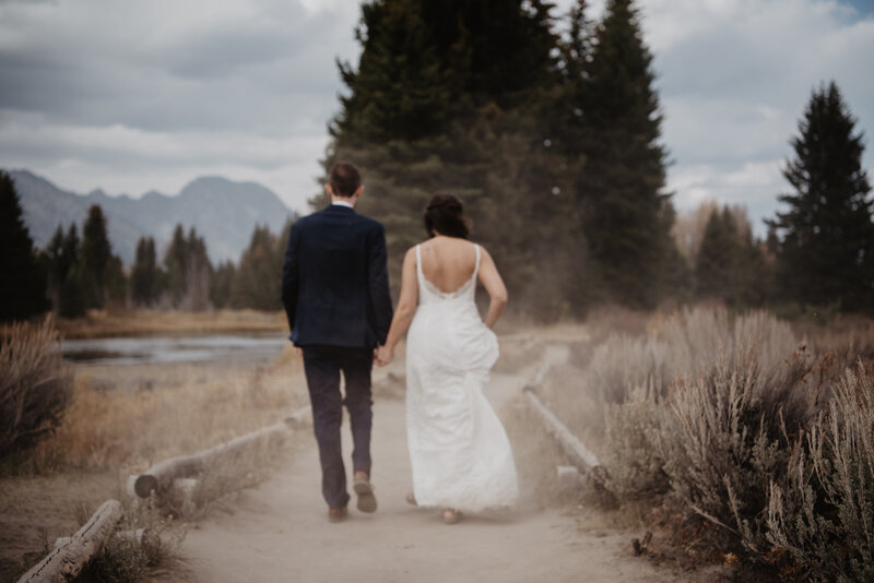 candid wedding pictures of wyoming elopement with bride and groom holding hands and walking on a dusty path together in the mountains for their fall wedding