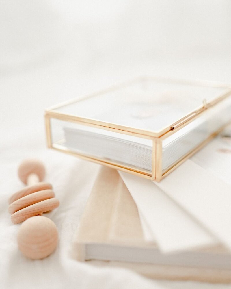 A close-up photo of a gold glass box with photo prints inside. A wooden baby rattle is placed by the box. The box sits on top of photo albums. 