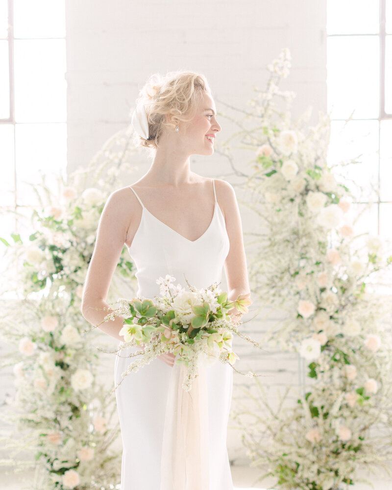 A smiling bride stands surrounded by airy, elegant white flowers at her wedding ceremony at L'Eloi. 