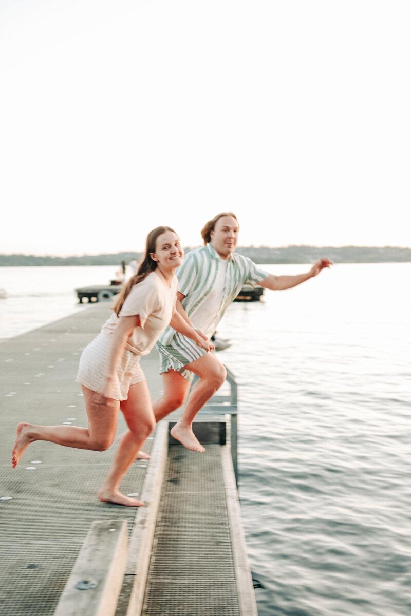 a couple hold hands as they jump off the dock into the water.