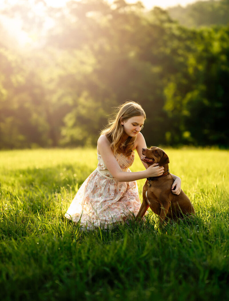 a Young girl in a floral sundress sits in a spot of sunlight with her vizla dog. The wind is blowing her hair. 